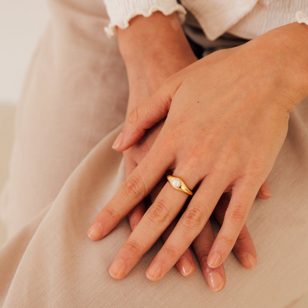 A close-up of hands wearing the Penelope Pearl Signet Ring, featuring a lustrous freshwater pearl set in gold, resting on beige fabric.