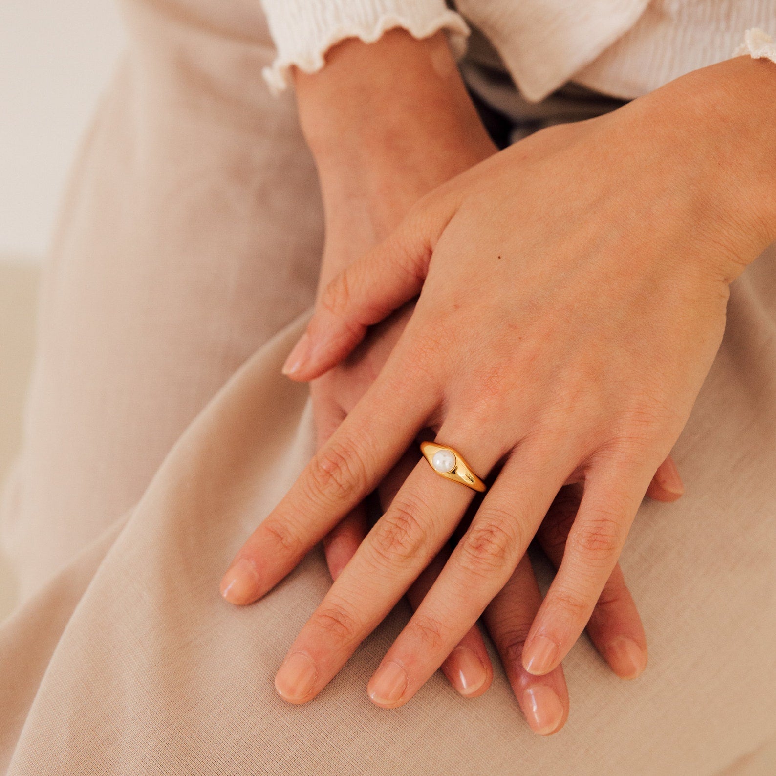 A close-up of hands wearing the Penelope Pearl Signet Ring, featuring a lustrous freshwater pearl set in gold, resting on beige fabric.