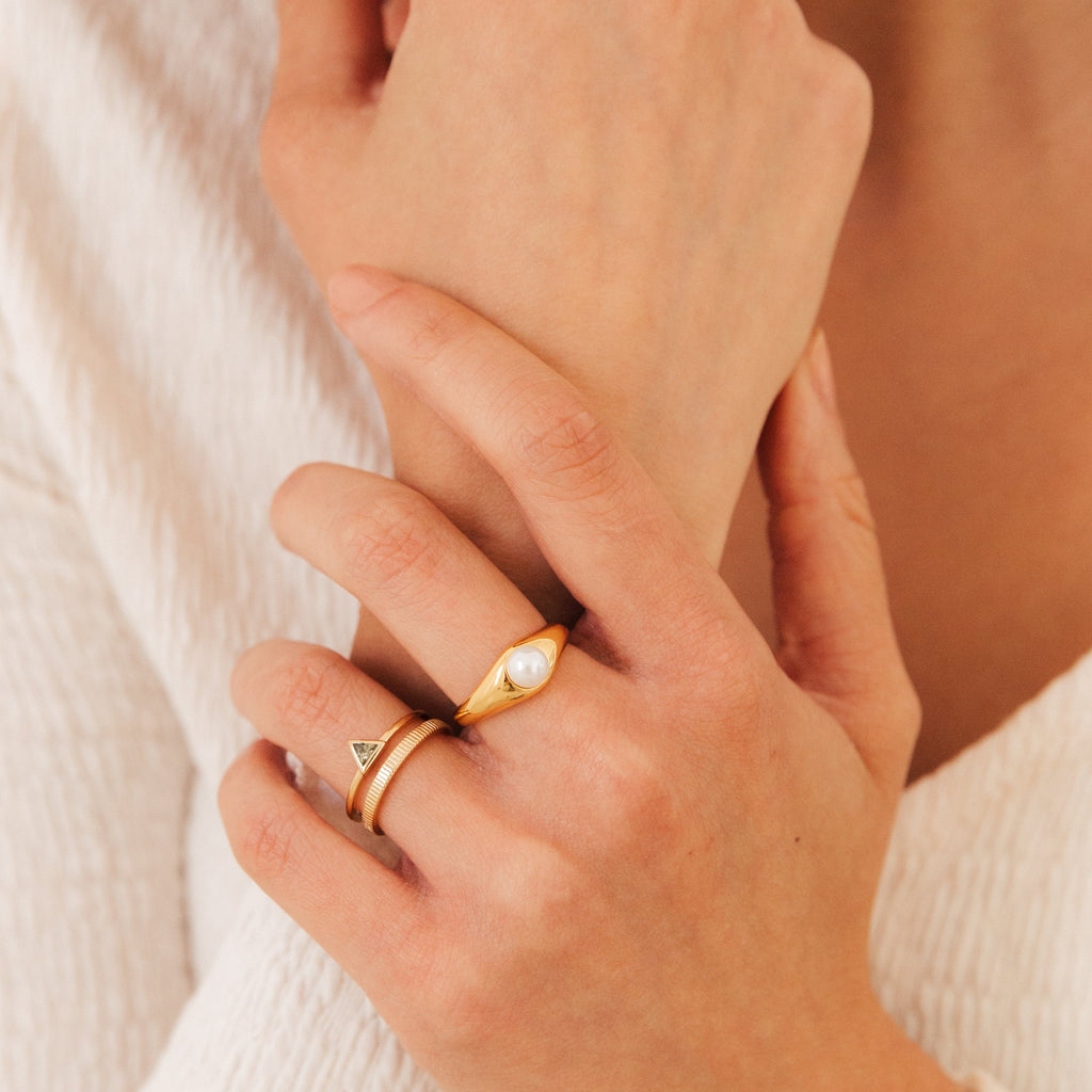A hand wears two gold rings—including the Penelope Pearl Signet Ring and a dainty triangle—against cream fabric, channeling vintage elegance.