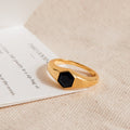 The Ember Signet Ring features a chunky gold design with a black hexagonal stone, displayed on textured beige fabric and a white card—an ideal stylish men's gift.