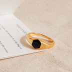The Ember Signet Ring features a chunky gold design with a black hexagonal stone, displayed on textured beige fabric and a white card—an ideal stylish men's gift.