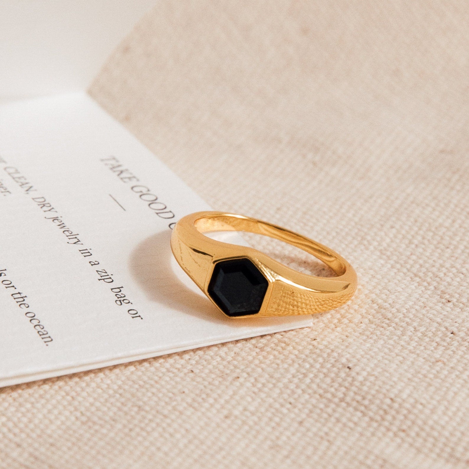 The Ember Signet Ring features a chunky gold design with a black hexagonal stone, displayed on textured beige fabric and a white card—an ideal stylish men's gift.