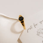 The Ember Signet Ring features a bold black hexagonal stone set in a gold chunky band, displayed on cream-colored paper with handwritten notes and sketches—a unique and striking men's gift.
