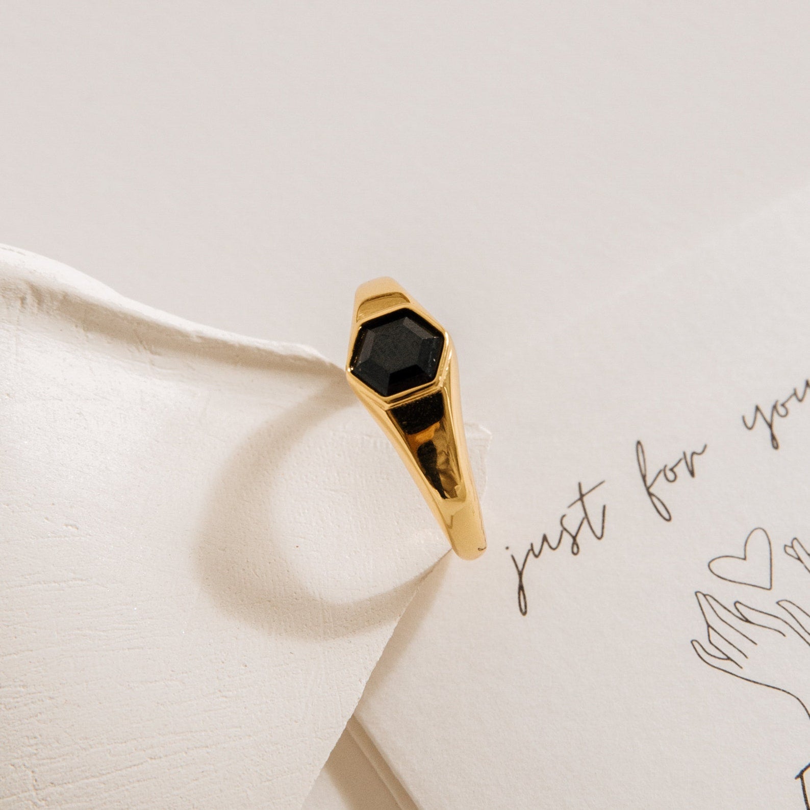 The Ember Signet Ring features a bold black hexagonal stone set in a gold chunky band, displayed on cream-colored paper with handwritten notes and sketches—a unique and striking men's gift.