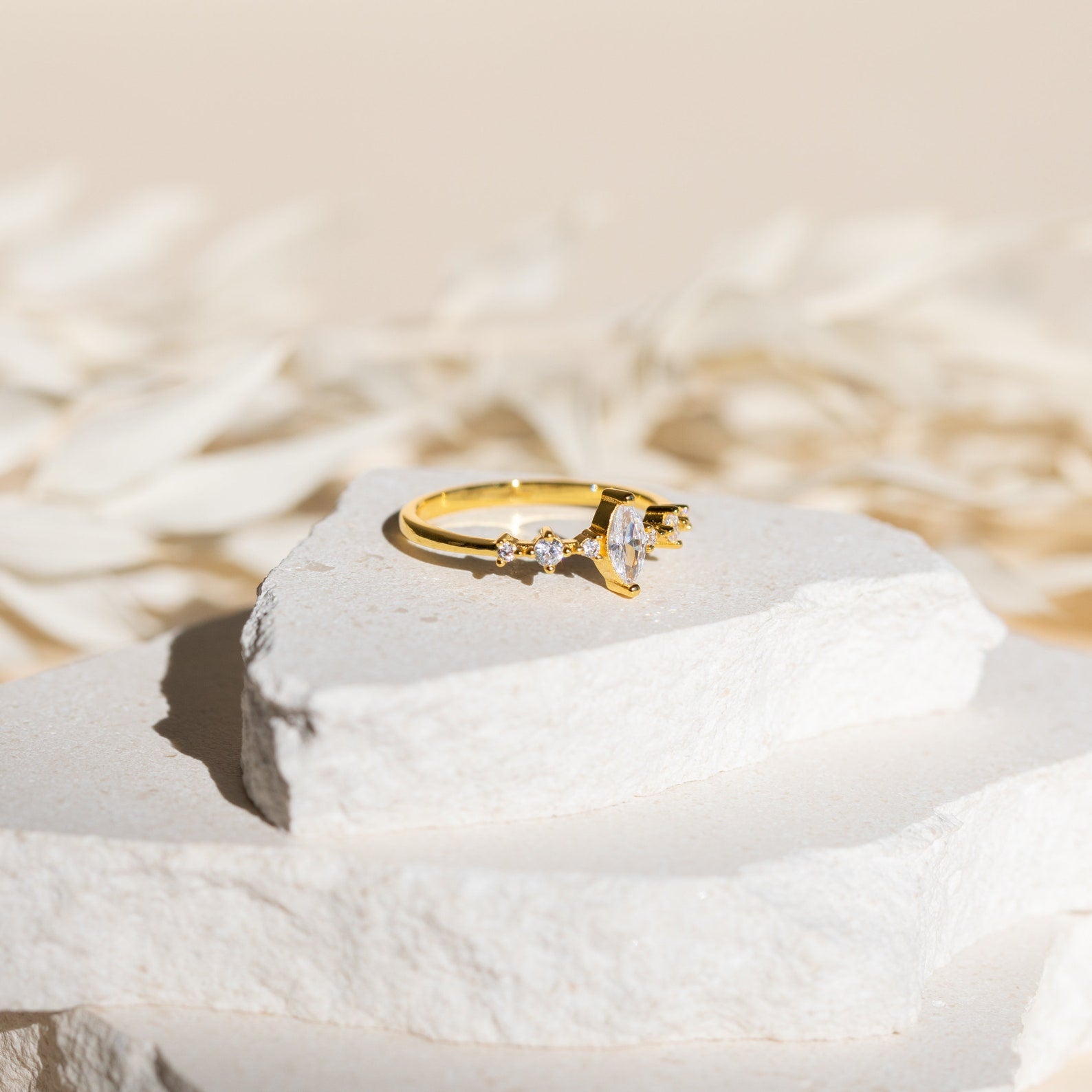 The Harlow Diamond Ring in 18K Gold, featuring a central oval stone and side diamonds, rests on stacked white stones against a soft beige background.