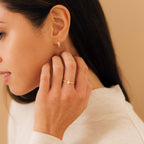 A woman in a cream top touches her face gently, wearing gold earrings and the Harlow Diamond Ring in 18K Gold.