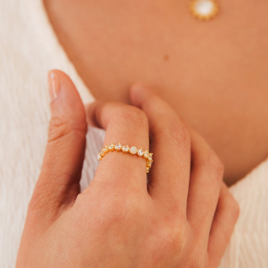 A hand displays the Mosaic Wave Eternity Ring—a gold band set with round gemstones—while a necklace appears blurred in the background.