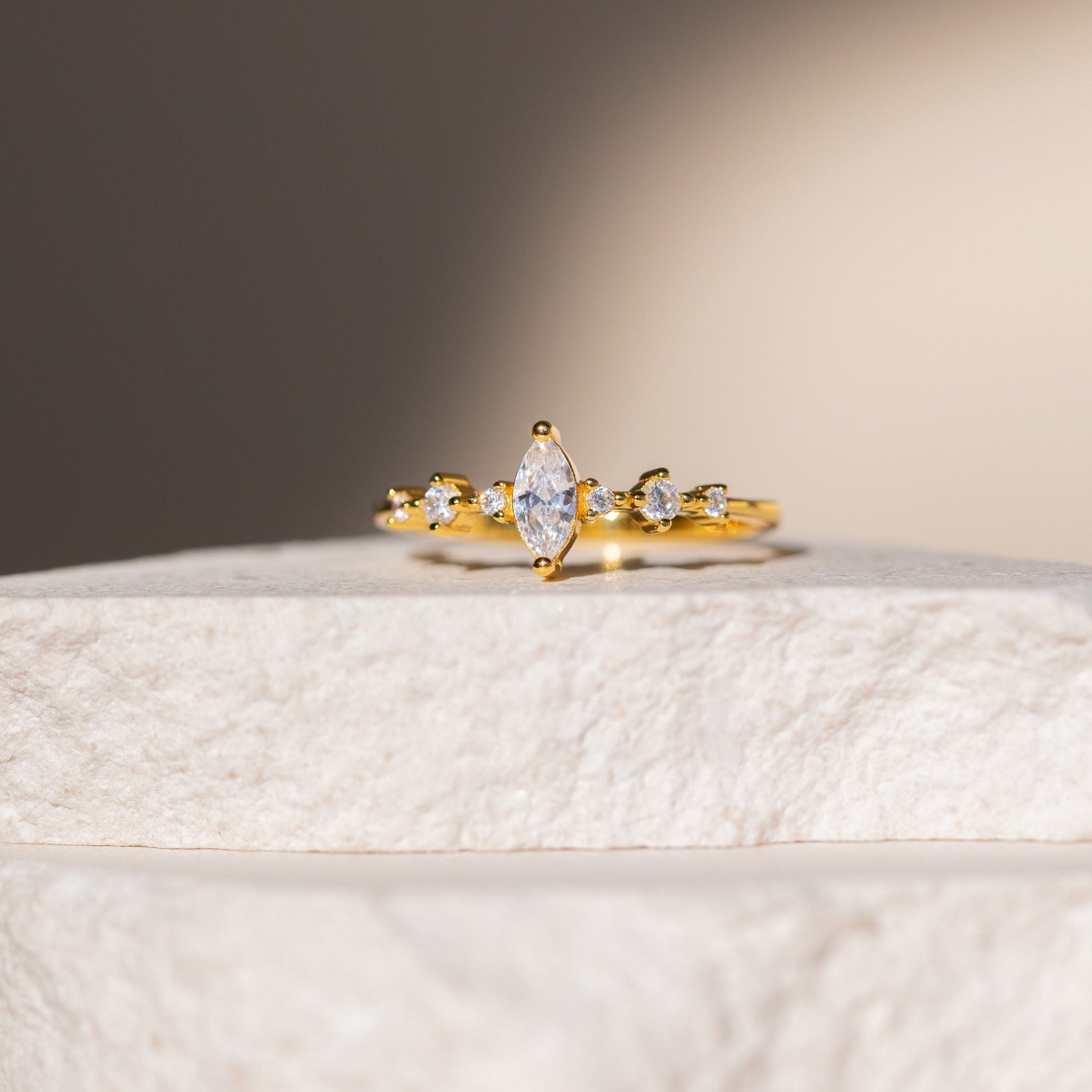 The Harlow Diamond Ring in 18K Gold, featuring a marquise diamond and round clear stones, is displayed on a textured white surface—offering timeless elegance ideal for an engagement ring.