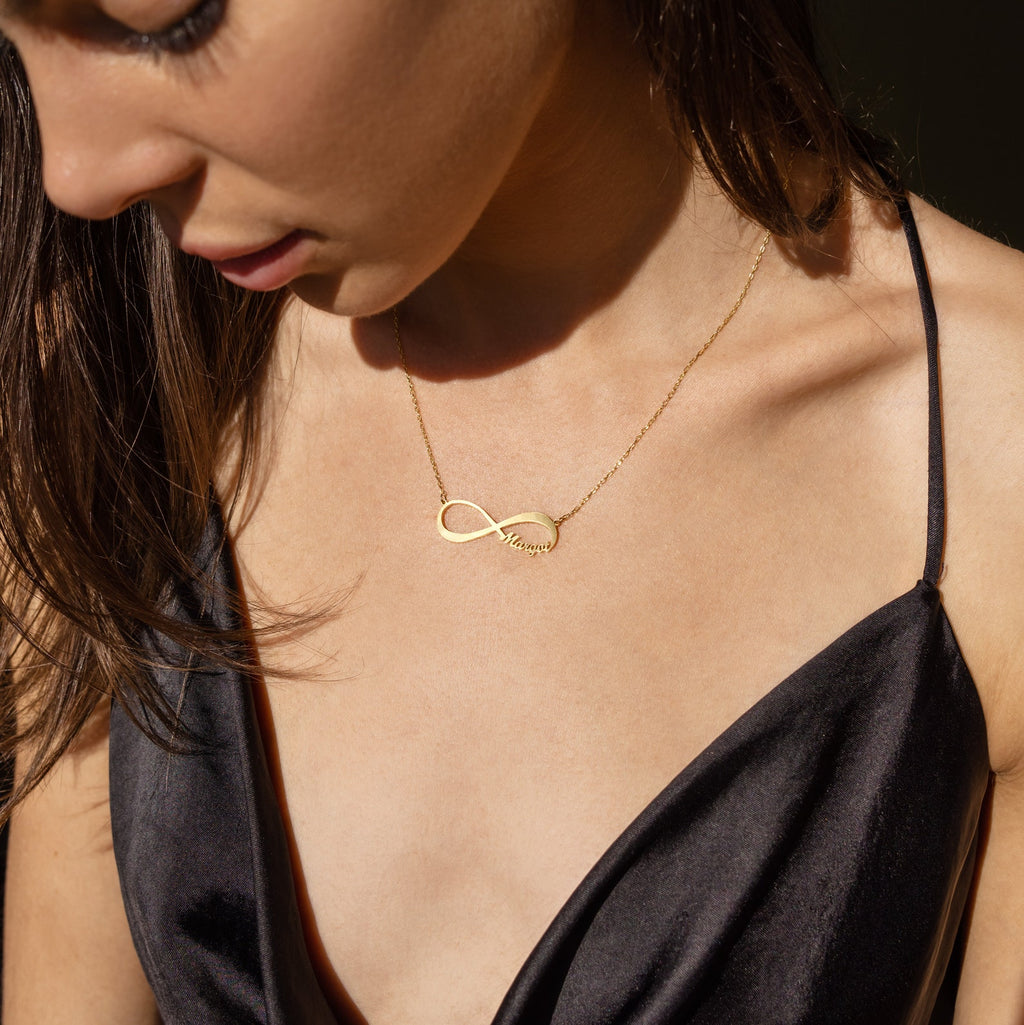 A woman in a black dress looks down in soft sunlight, wearing the Infinity Name Necklace.