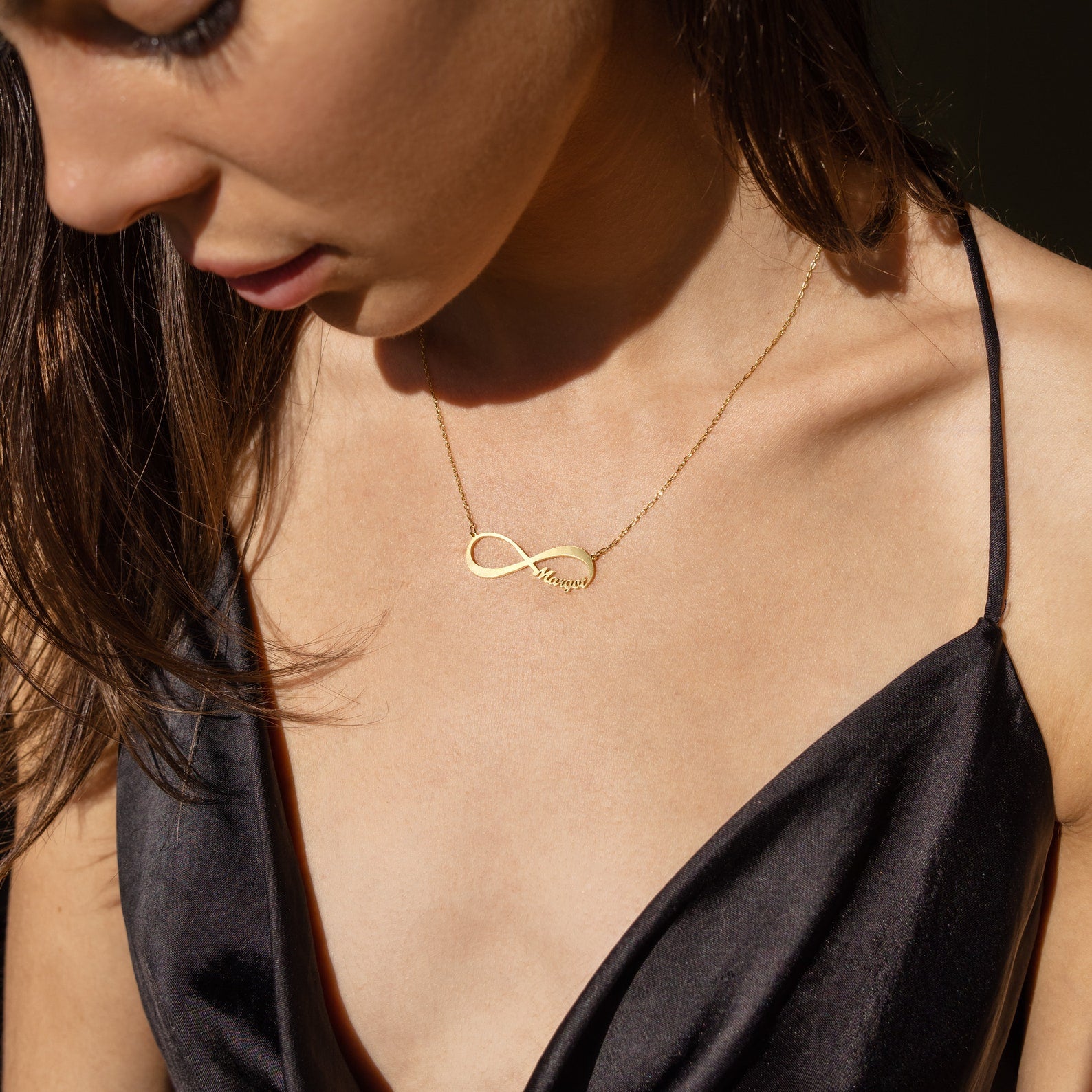 A woman in a black dress looks down in soft sunlight, wearing the Infinity Name Necklace.