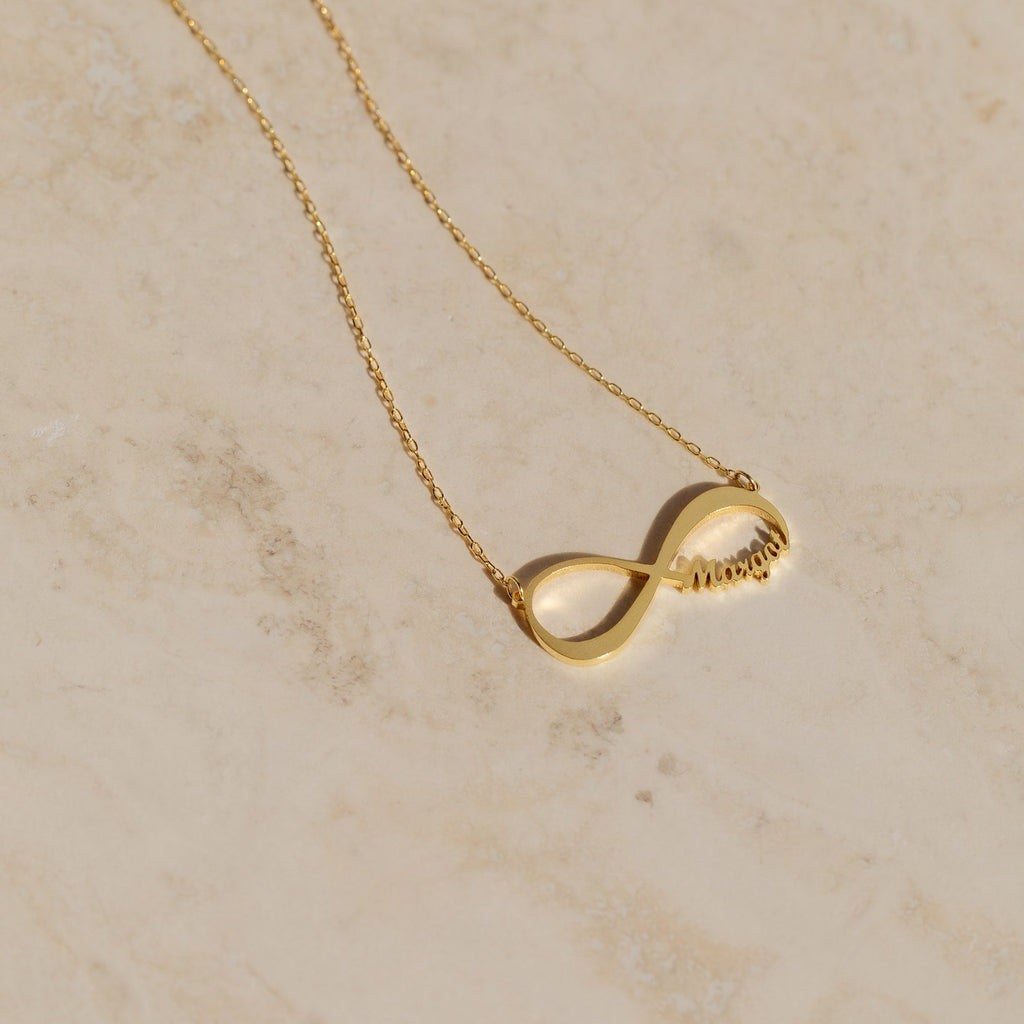 The Infinity Name Necklace features a delicate chain with your name in cursive, displayed on a light marble surface—an ideal personalized or anniversary gift for someone special.