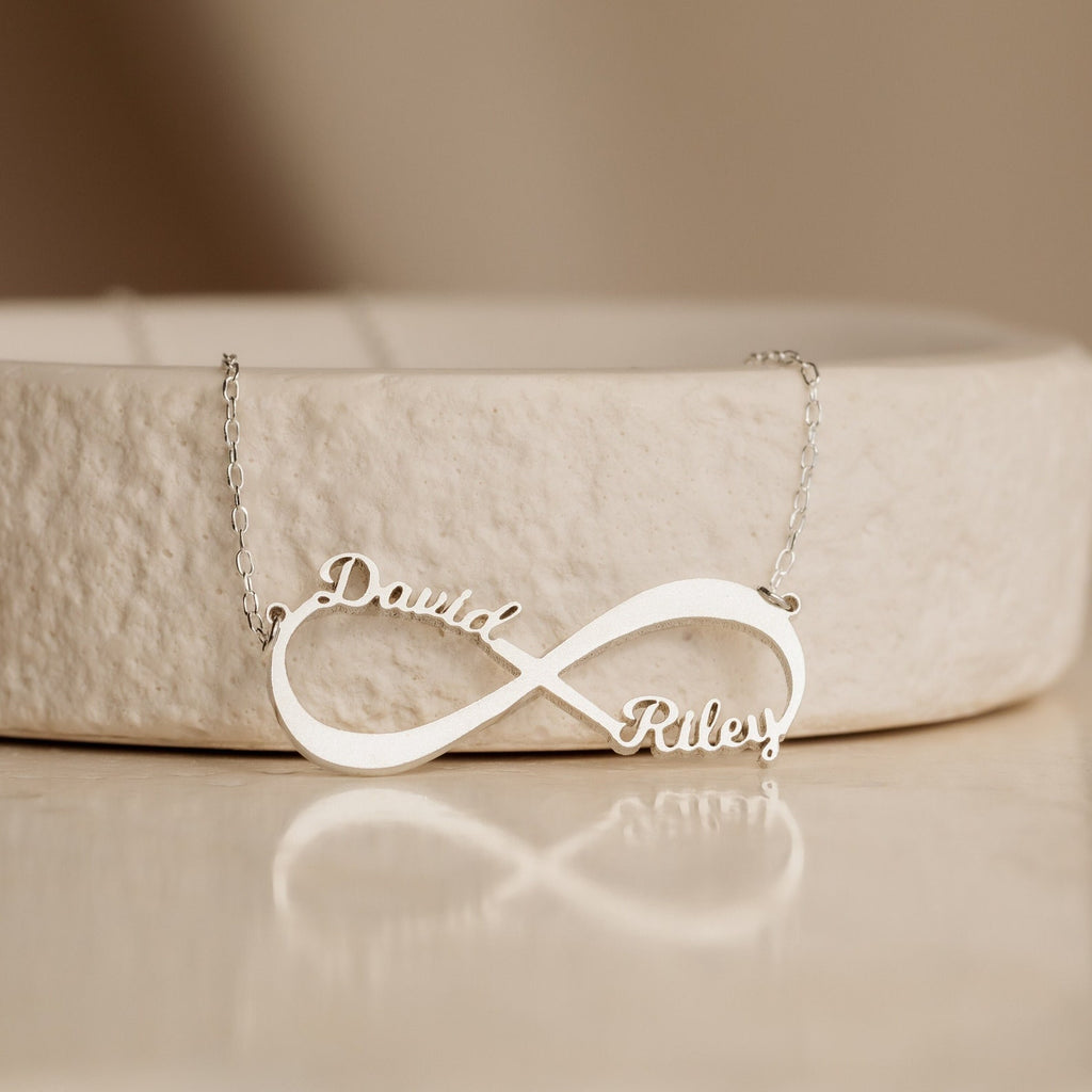 Infinity Name Necklace customized with the names “David” and “Riley,” displayed on a light beige textured surface. A heartfelt anniversary gift to treasure always.