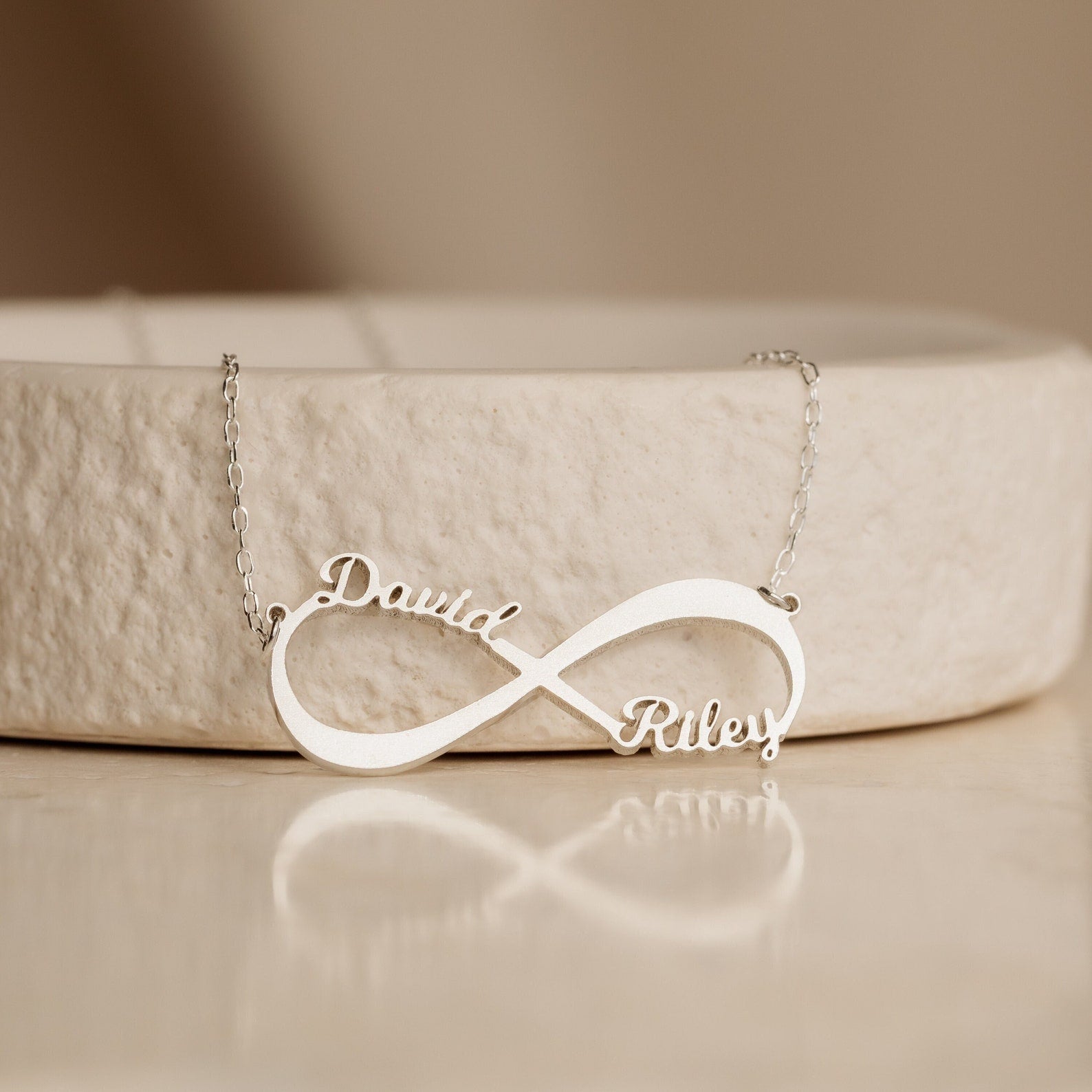 Infinity Name Necklace customized with the names “David” and “Riley,” displayed on a light beige textured surface. A heartfelt anniversary gift to treasure always.
