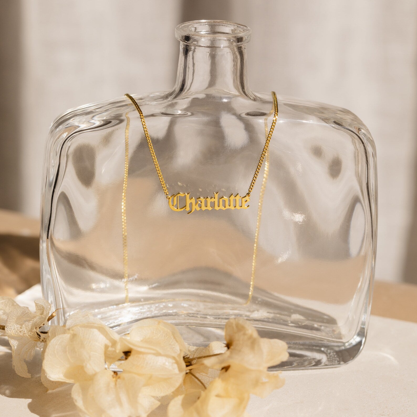 The Old English Name Necklace in Curb Chain rests on a clear glass bottle, showcasing a personalized charm, with beige flowers in the foreground.