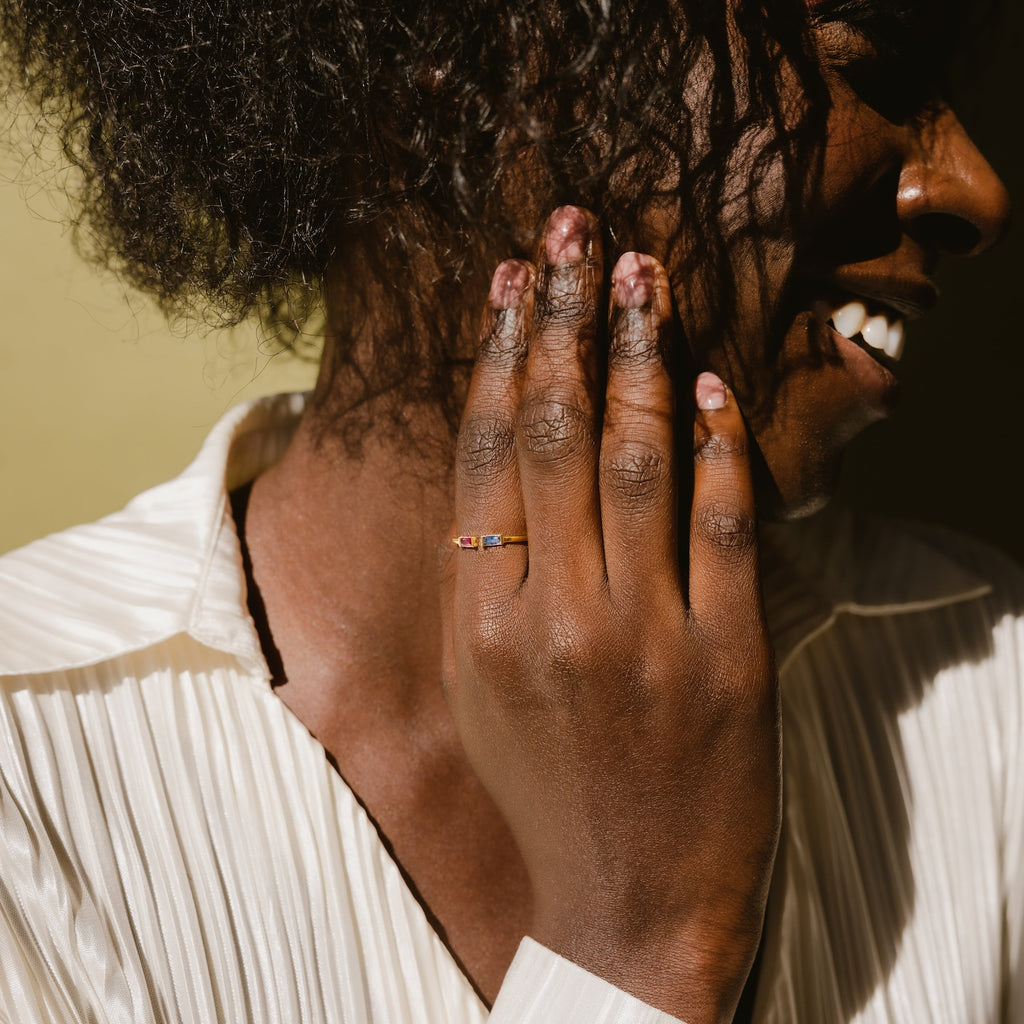 A smiling person touches their cheek, wearing the Duo Baguette Birthstone Ring with personalized gemstones, paired with a light blouse.
