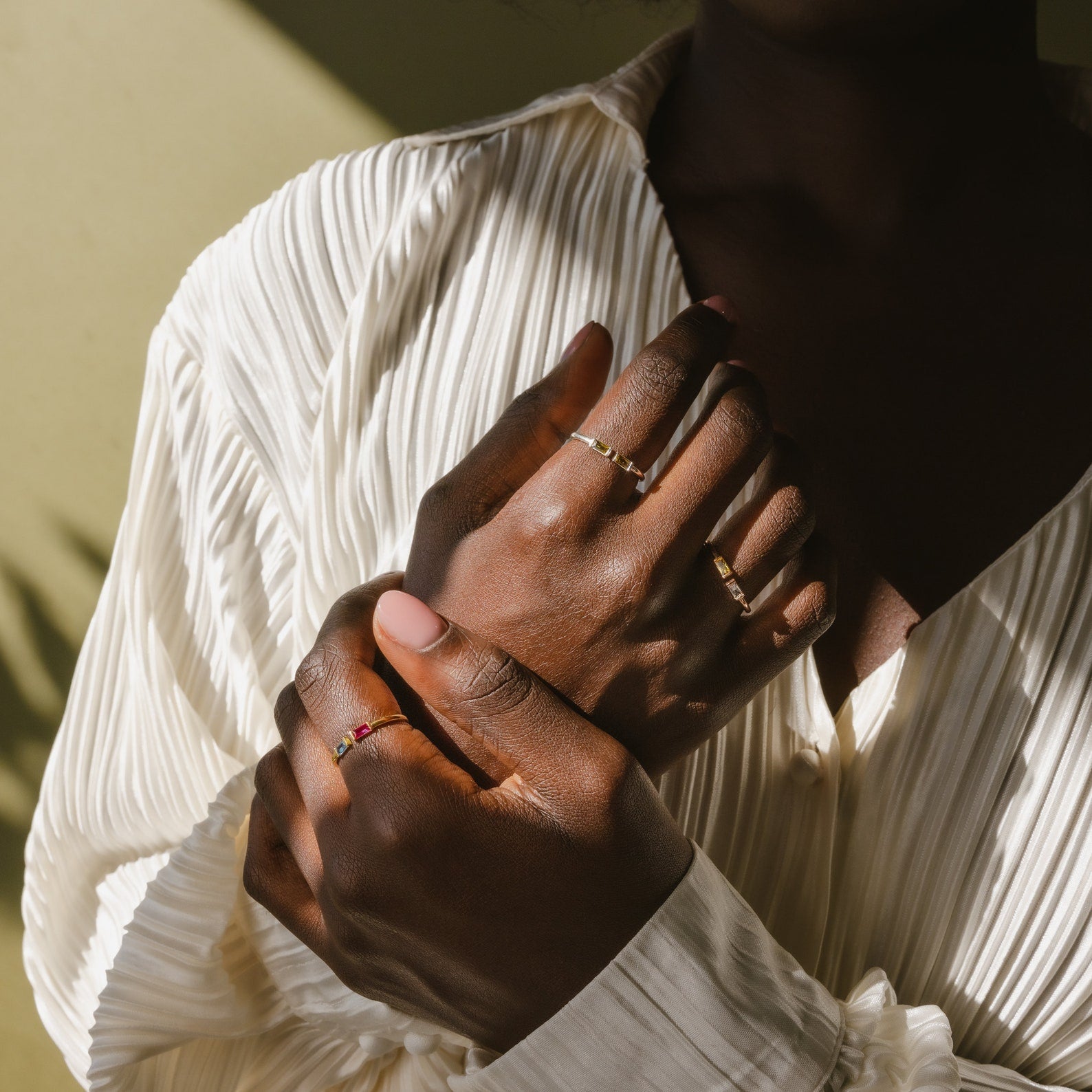 A person in a white pleated shirt displays hands adorned with delicate rings, including the Duo Baguette Birthstone Ring—an elegant, personalized piece that gleams beautifully in natural light.
