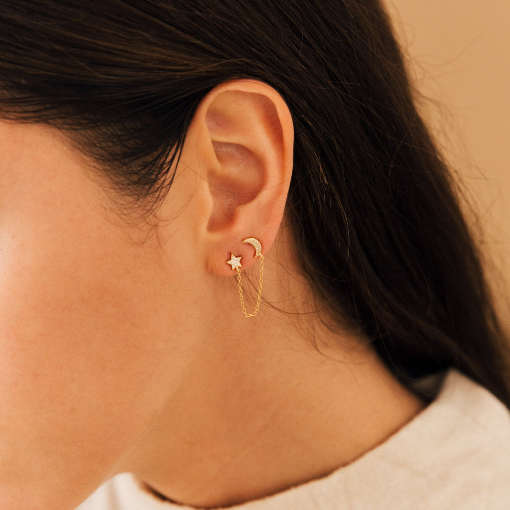 Close-up of a woman's ear wearing Luna Moon & Star Chain Earrings, showcasing a delicate gold chain connecting her cartilage piercing, adorned with moon and star studs.