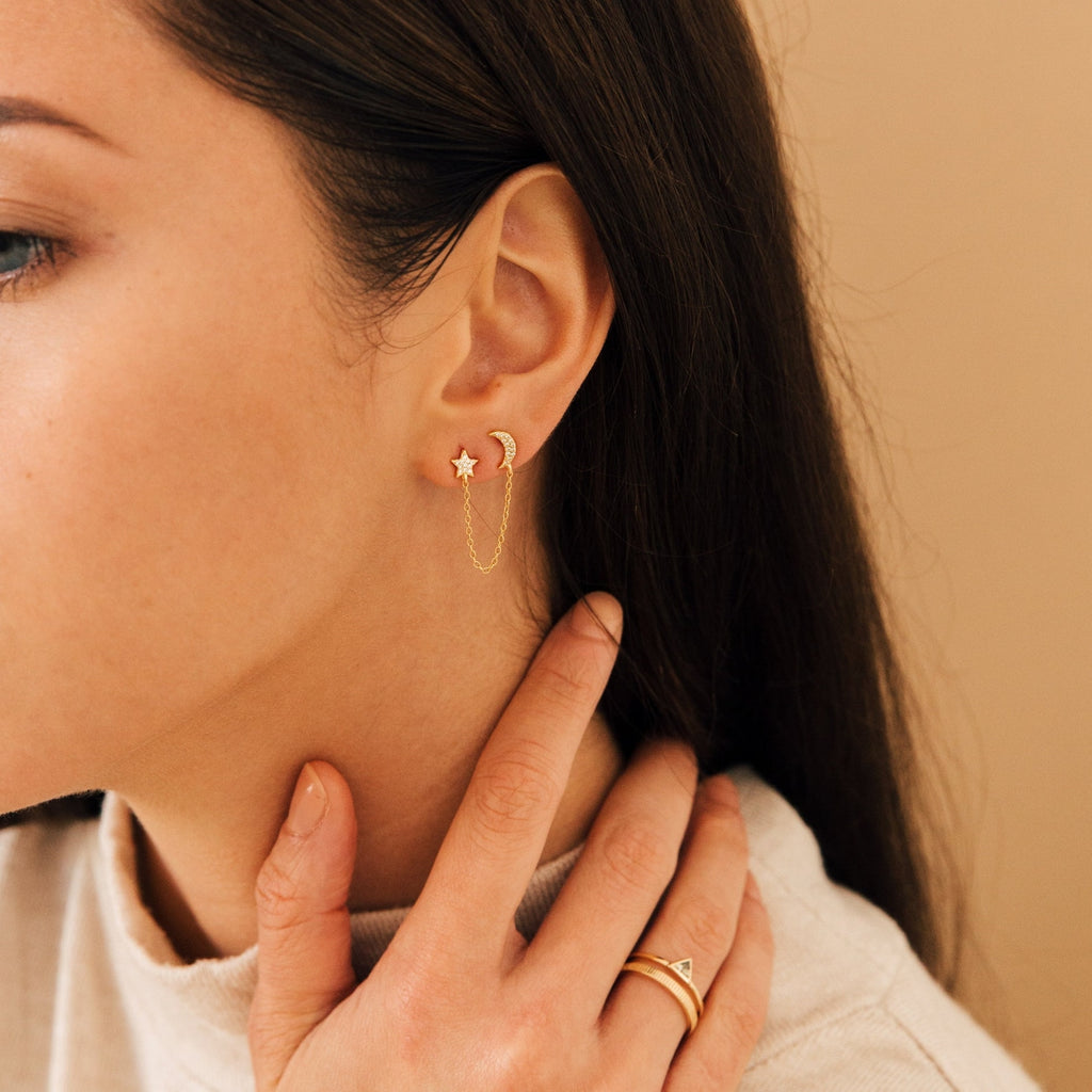 A woman wearing Luna Moon & Star Chain Earrings with a delicate cartilage piercing, touching her neck and displaying a gold ring on her finger.
