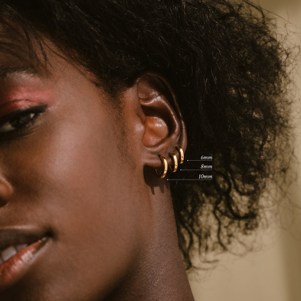 Close-up of a person with short curly hair and pink eyeshadow, wearing the Editor Huggie Earrings Set—three minimalist gold hoops in 6mm, 8mm, and 10mm. Also available in sterling silver.