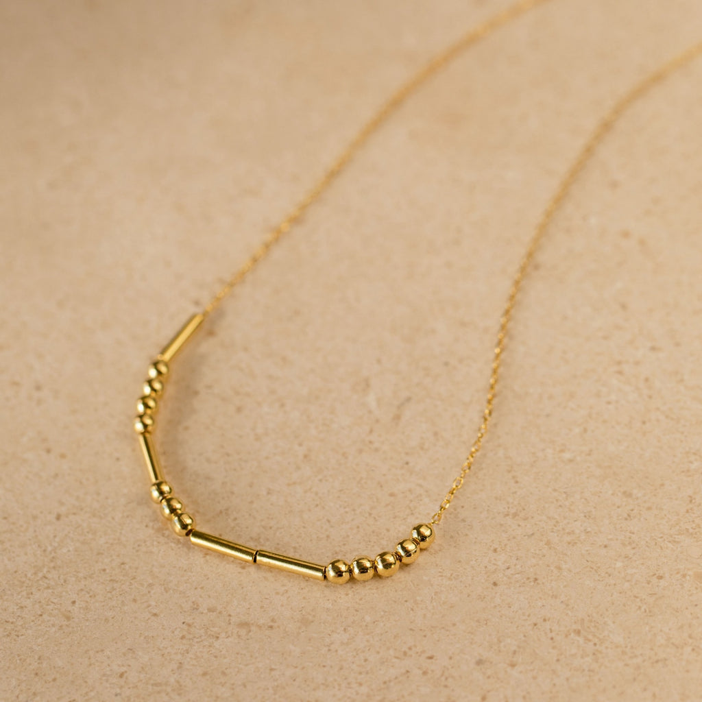 Morse Code Necklace with small round and tube-shaped beads on a delicate chain, shown on a beige surface—makes a unique, personalized gift.