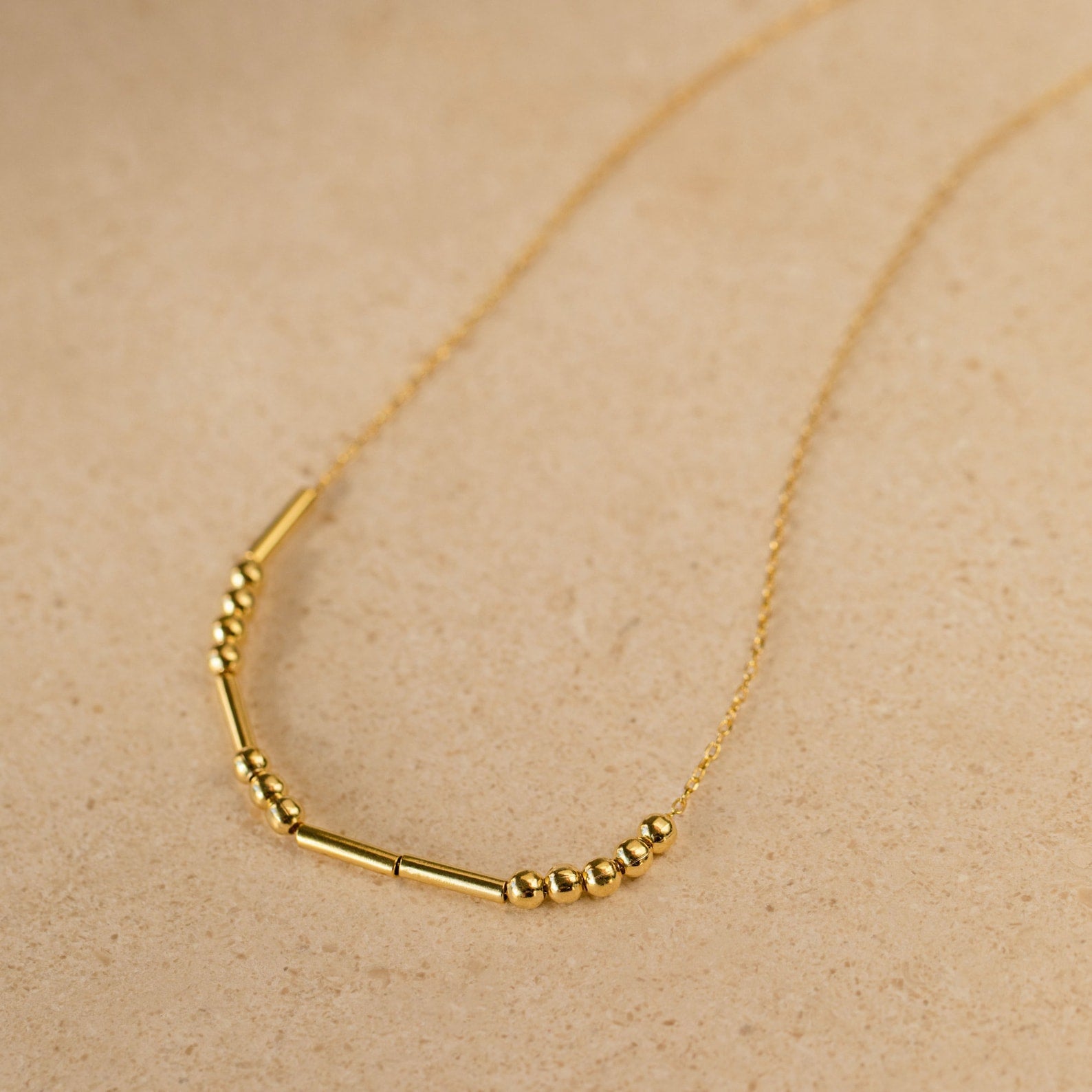 Morse Code Necklace with small round and tube-shaped beads on a delicate chain, shown on a beige surface—makes a unique, personalized gift.