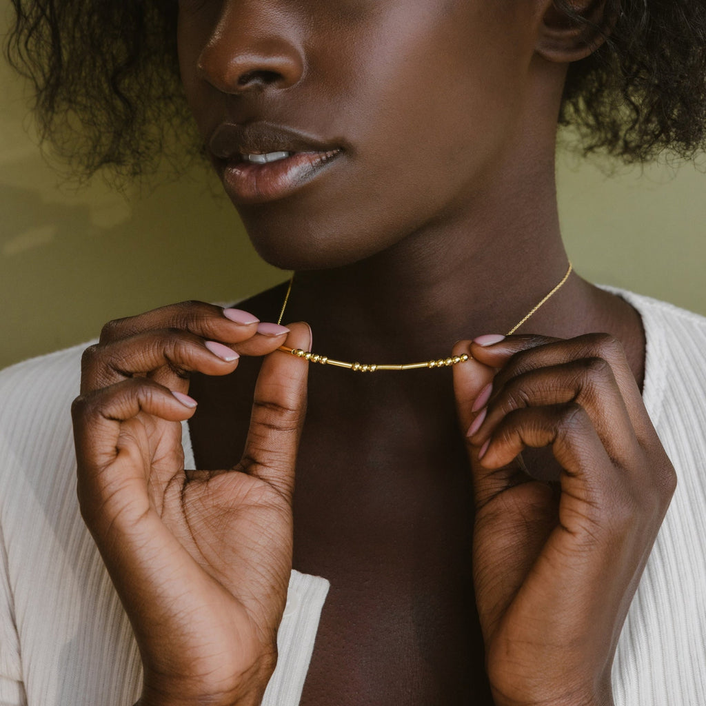 A woman with dark skin wears a white top and holds the Morse Code Necklace near her neck—this unique piece makes a thoughtful, personalized gift.