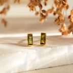 Baguette Birthstone Studs: Rectangular green gemstone earrings in gold settings rest on stone, with dried flowers behind—chic and personalized gift-ready birthstone studs.