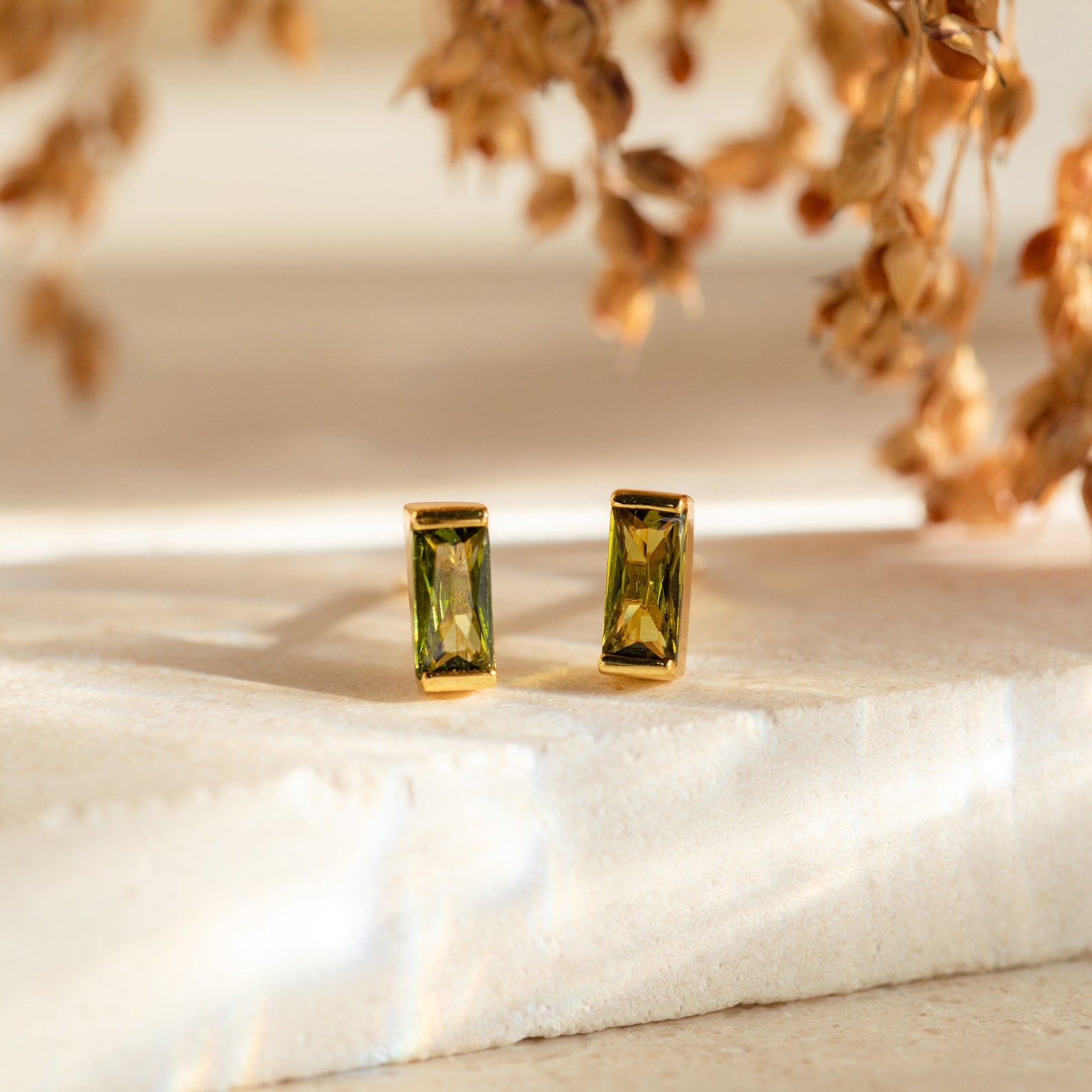 Baguette Birthstone Studs: Rectangular green gemstone earrings in gold settings rest on stone, with dried flowers behind—chic and personalized gift-ready birthstone studs.