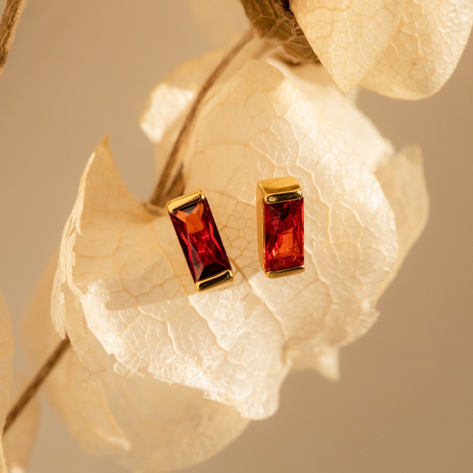 Baguette Birthstone Studs featuring rectangular red gemstones, displayed on a white dried leaf—ideal as birthstone earrings or personalized gifts.