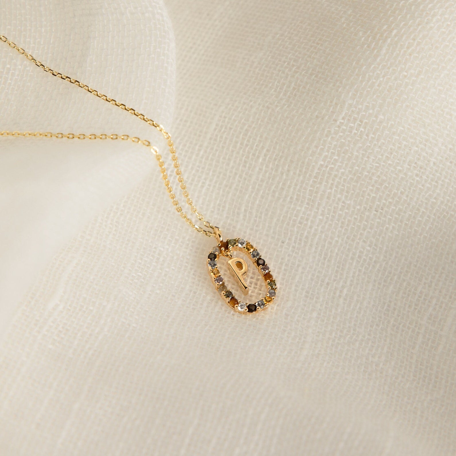 Rainbow Initial Necklace featuring a gold pendant with the letter P, bordered by small gemstones, displayed on white fabric—a stunning personalized accessory that adds a unique touch to any look.