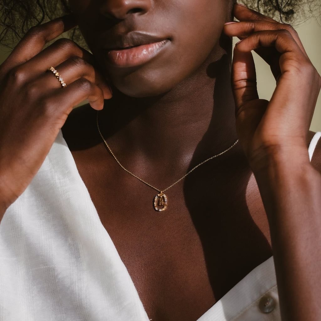 Woman in a white top wearing the Rainbow Initial Necklace and a gold ring, with her hands near her neck.