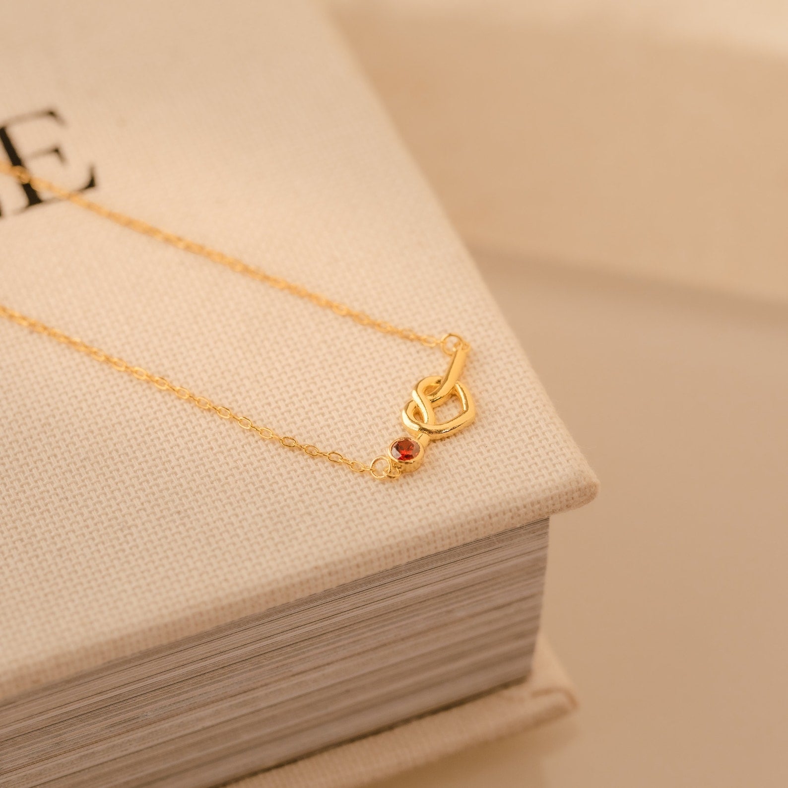 The Love Knot Birthstone Necklace, featuring a gold chain with a small red gemstone and heart-shaped clasp, lies on a closed beige book.