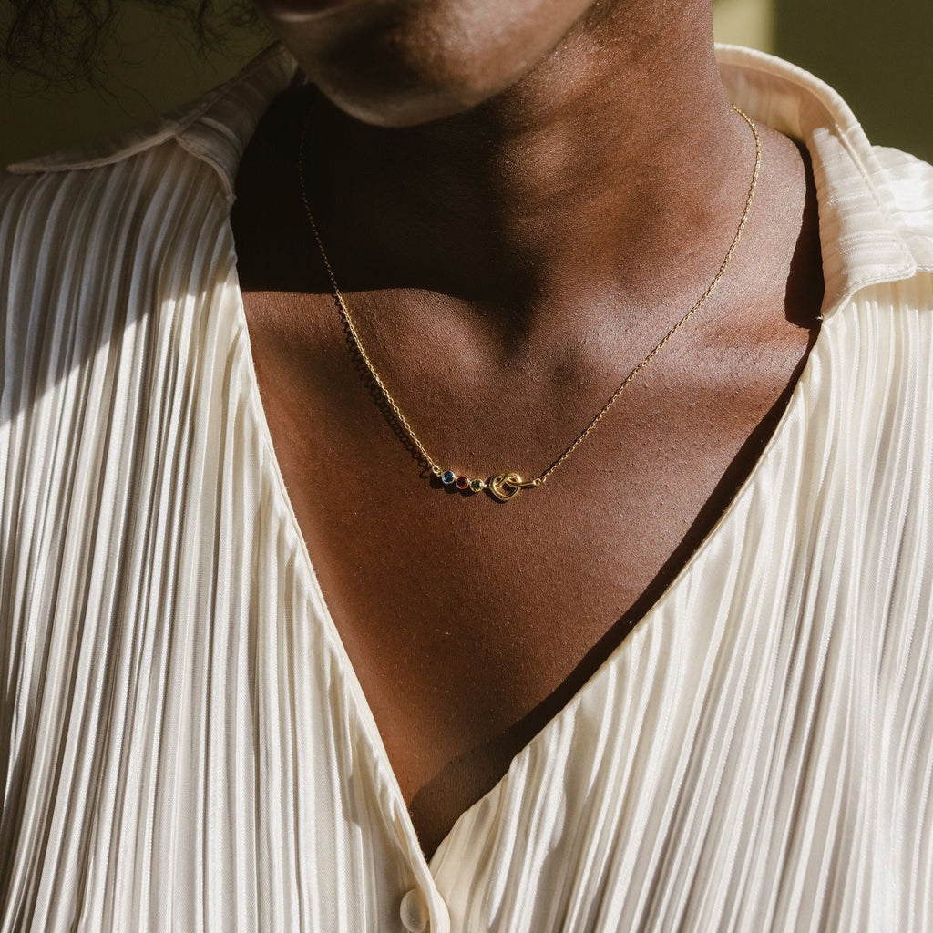 A person wearing a pleated cream blouse and a gold Love Knot Birthstone Necklace featuring colorful beads and a moon charm.