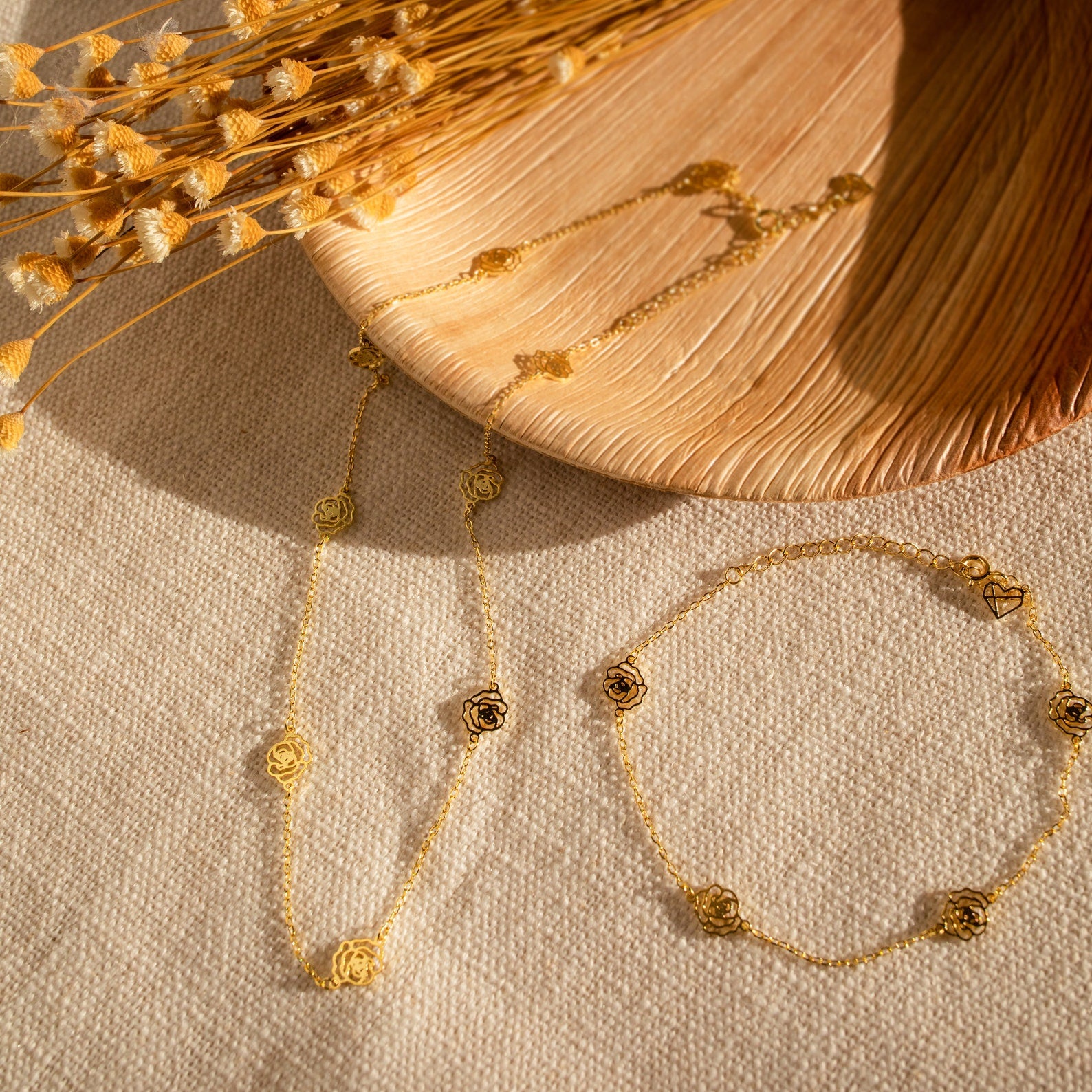 The gold Flower Station Necklace rests on textured beige fabric with dried flowers and a wooden plate—a perfect piece of floral jewelry and a thoughtful gift for her.