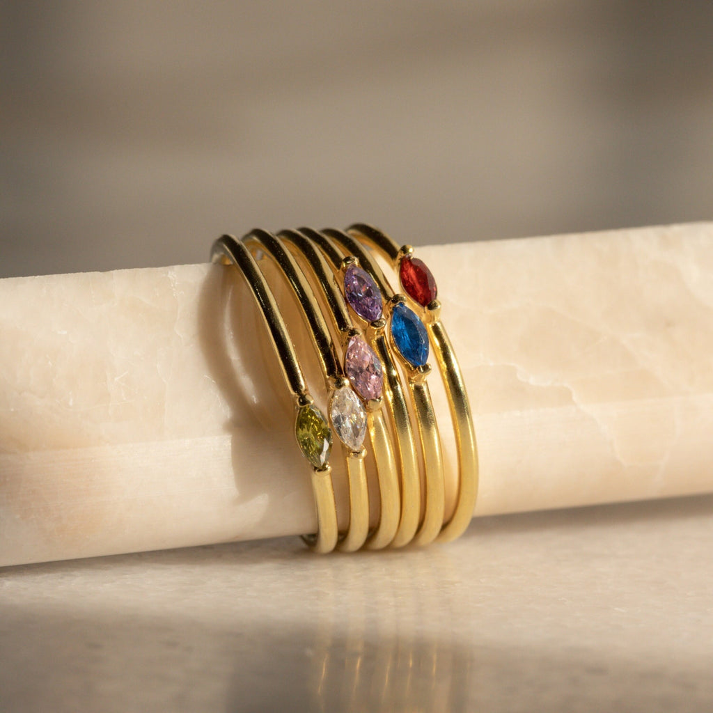 Six gold rings, featuring the Marquise Birthstone Ring, are stacked on a marble rod and bathed in soft natural light.