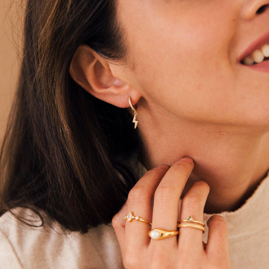 A woman smiles softly, showing off her trendy jewelry with a hand near her ear while wearing Striker Pave Huggies and rings.