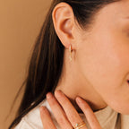Close-up of a woman wearing trendy gold lightning bolt earrings, Striker Pave Huggies, and gold rings while gently touching her neck.