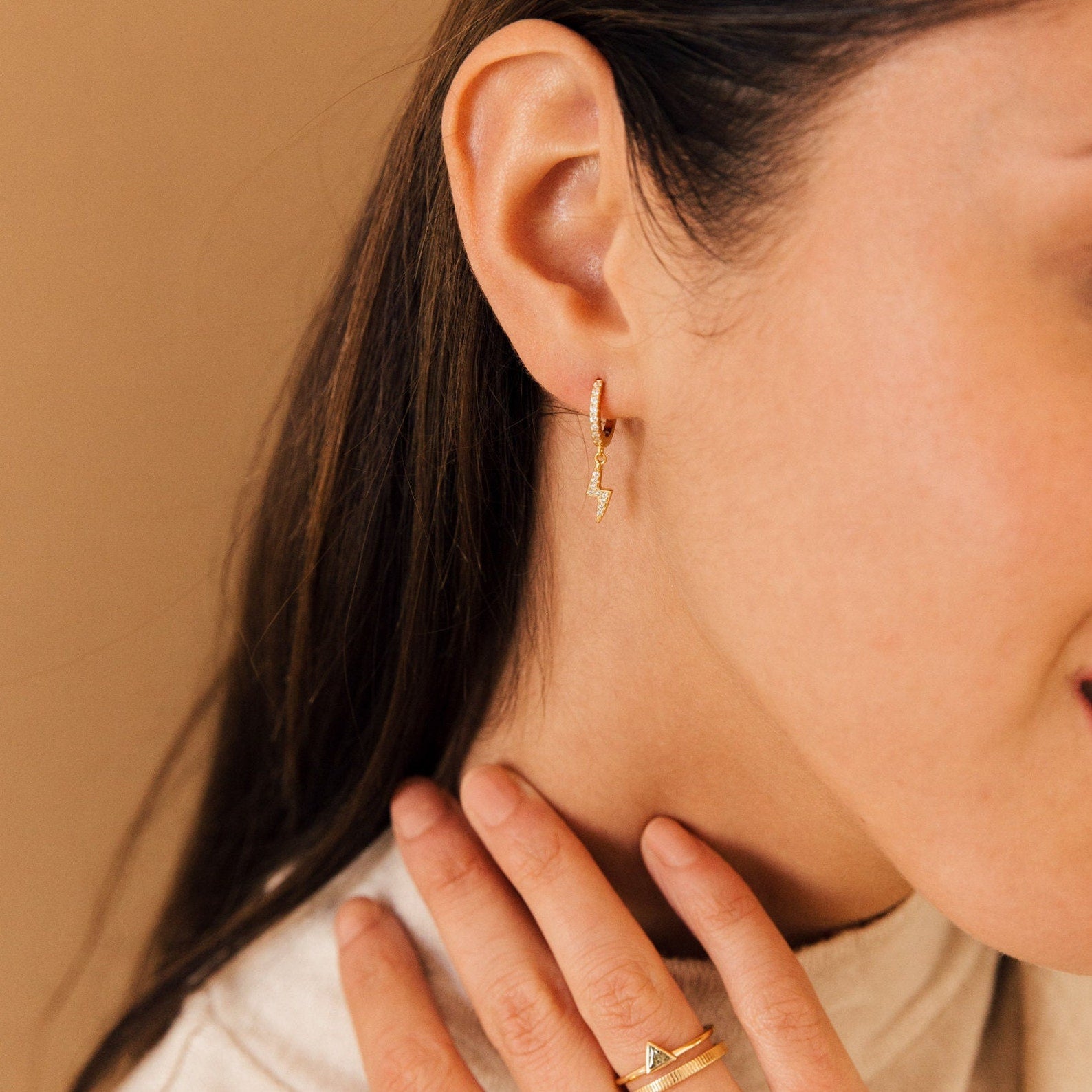 Close-up of a woman wearing trendy gold lightning bolt earrings, Striker Pave Huggies, and gold rings while gently touching her neck.