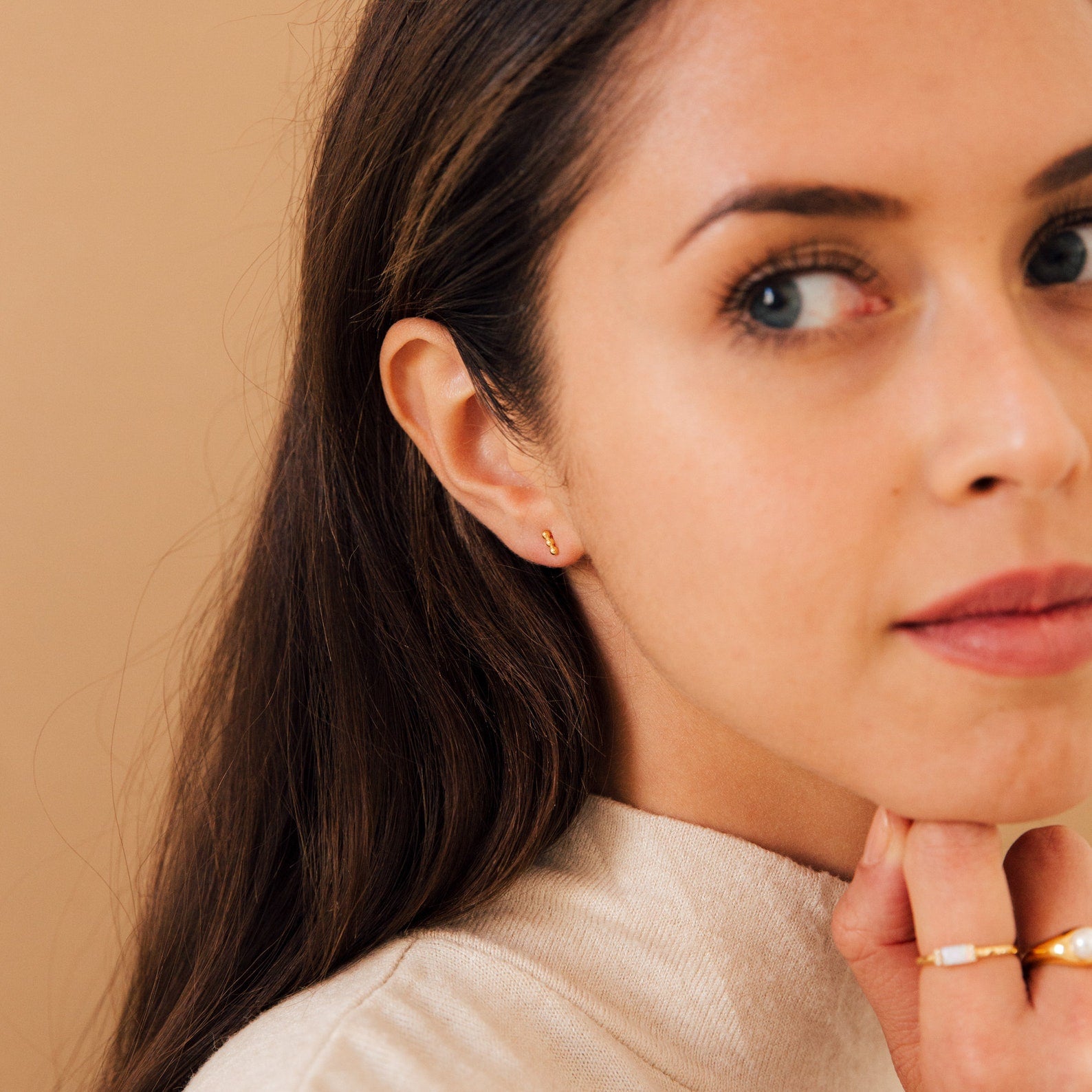 Woman with long brown hair, wearing gold rings and Trio Beaded Bar Studs, looks slightly to the side—perfect inspiration for everyday wear.