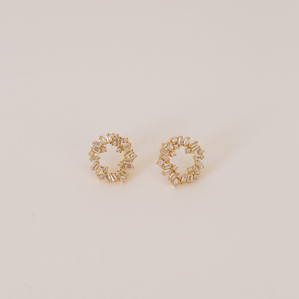 The Reina Cluster Studs are gold circular earrings featuring baguette-cut stones in a wreath-like pattern, making them the perfect elegant bridal accessory for your special day.