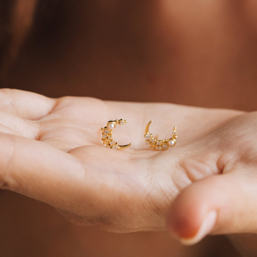 A hand holds two Eclipse Moon Studs—gold crescent moon earrings adorned with pearls and tiny beads.