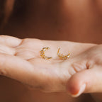 A hand holds two Eclipse Moon Studs—gold crescent moon earrings adorned with pearls and tiny beads.