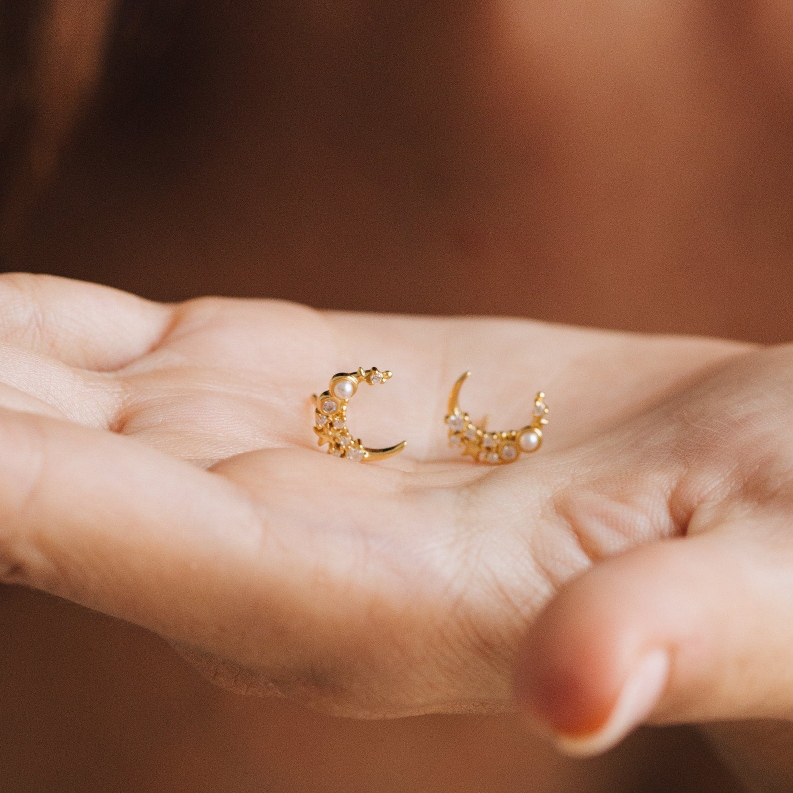A hand holds two Eclipse Moon Studs—gold crescent moon earrings adorned with pearls and tiny beads.