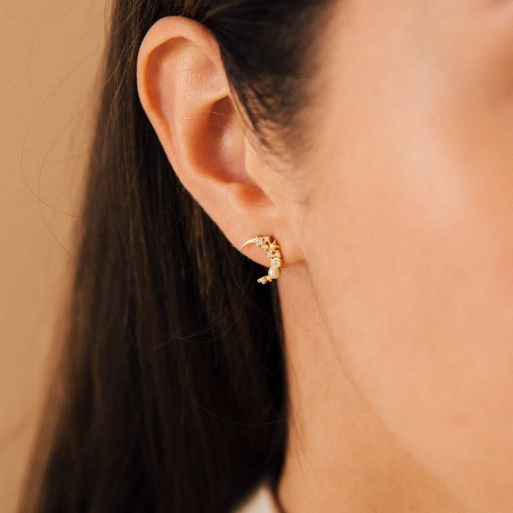 Close-up of a woman's ear wearing Eclipse Moon Studs—delicate, gold, and curved earrings adorned with tiny stones that sparkle like stars.