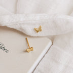 The Emilia Butterfly Studs, elegant gold earrings, rest on soft white fabric beside a closed book—perfect for fans of dainty jewelry.