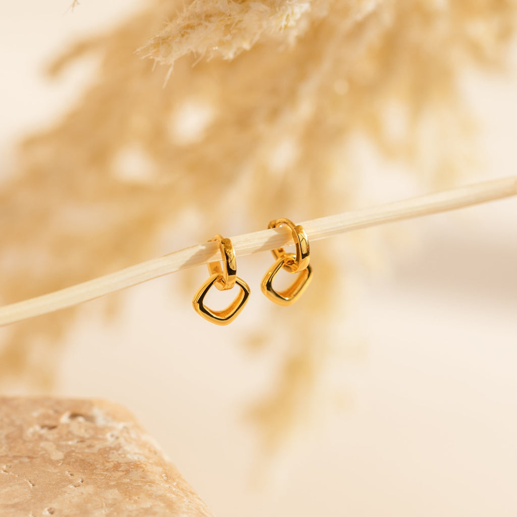 Reagan Duo Huggies dangle elegantly from a slender branch, featuring gold geometric hoops that shine against a subtle, neutral backdrop.