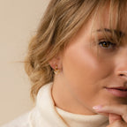 Woman with blonde hair wearing a cream turtleneck and chic earrings touches her chin, showcasing the Reagan Duo Huggies for a stylish, modern look.