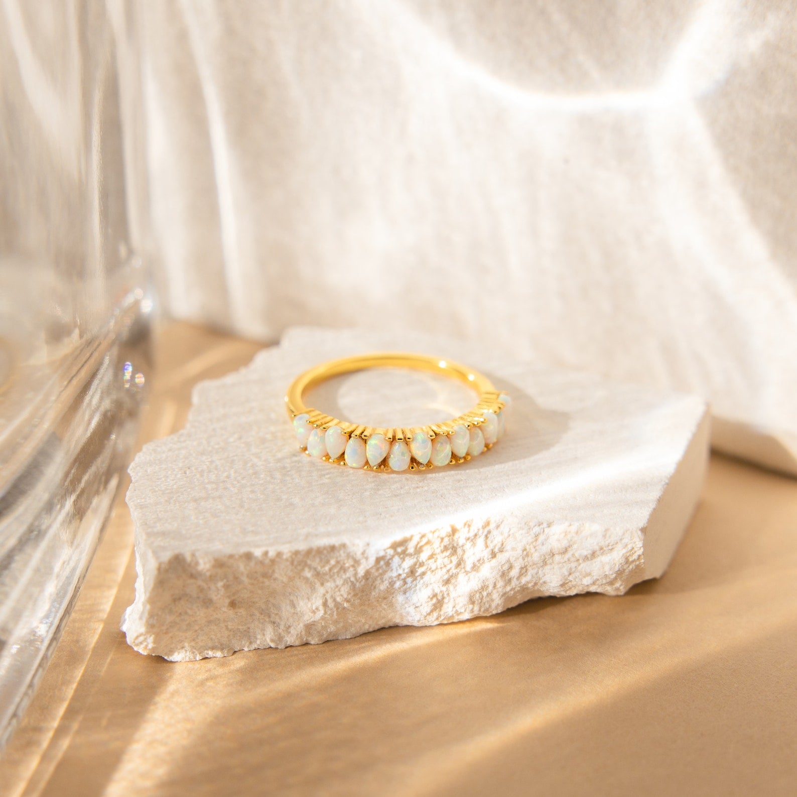 The Opal Drop Eternity Ring, a vintage-inspired gold piece with shimmering white gems, is displayed on a rough white stone in soft natural light against a neutral background.