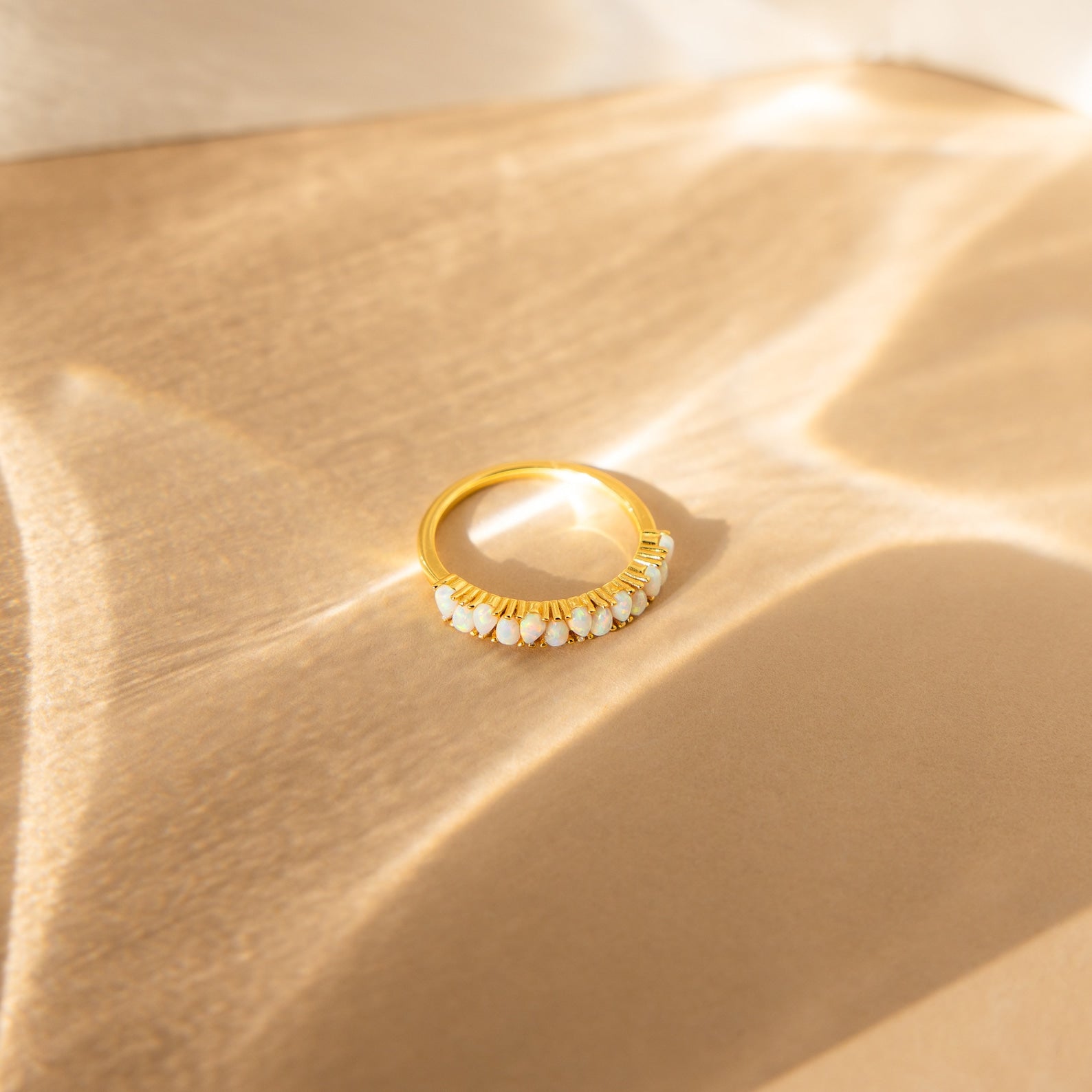 The Opal Drop Eternity Ring, a vintage-inspired gold band adorned with opals and small white stones, rests on a beige surface illuminated by gentle natural sunlight.