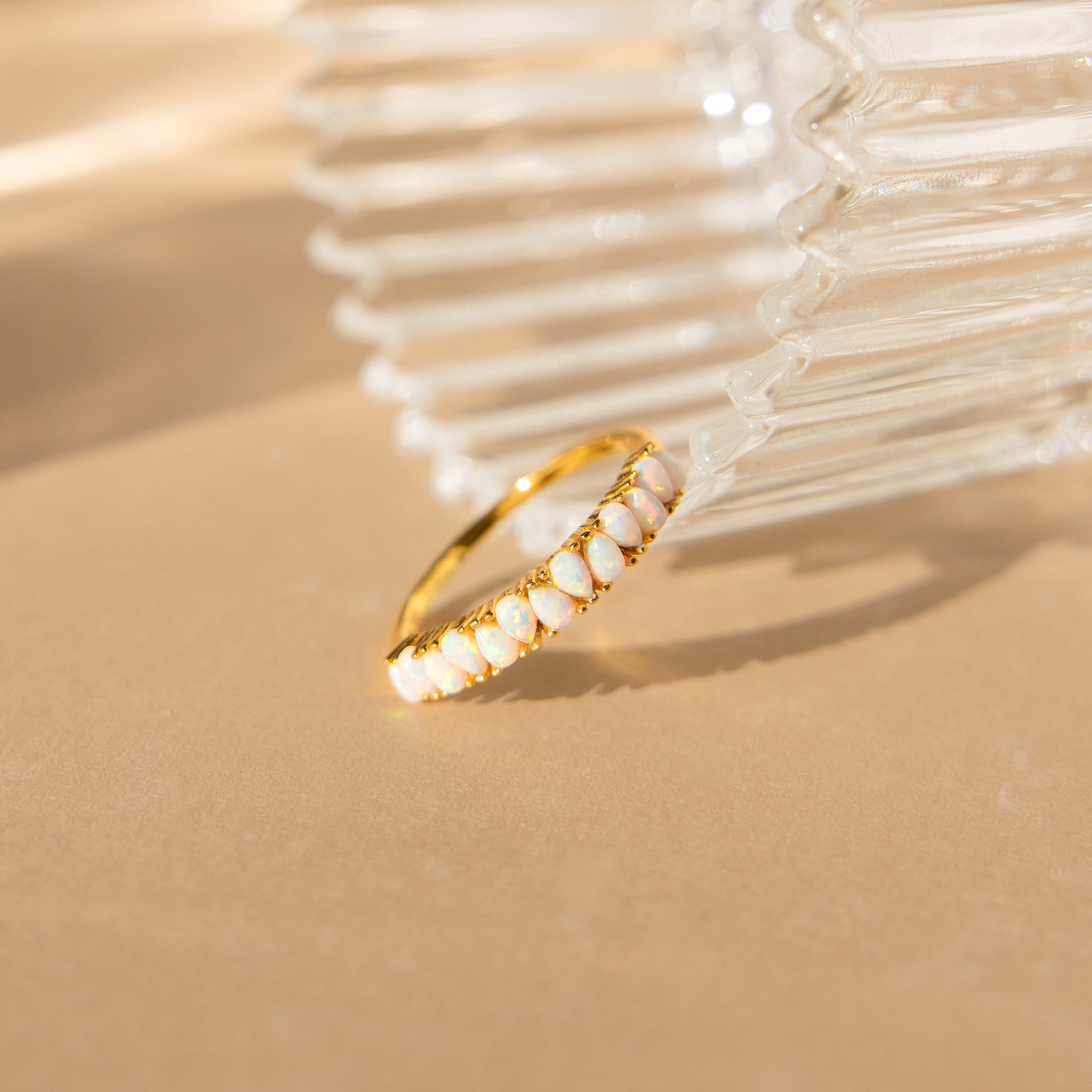 The Opal Drop Eternity Ring, a vintage-inspired piece with opal gemstones, is displayed against a ribbed glass accent on a beige surface.